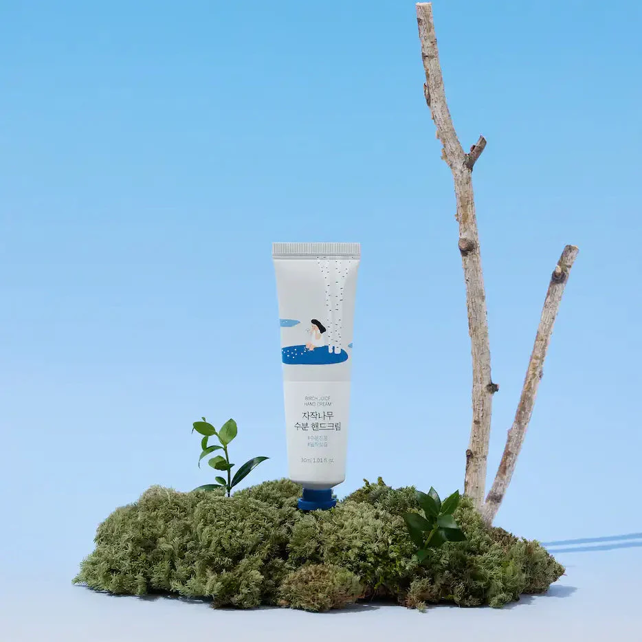 A tube of birch hand cream surrounded by green moss and small branches against a blue background.