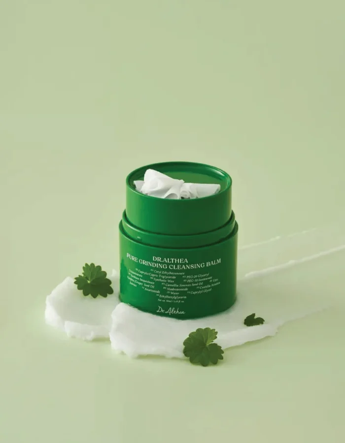 Green cleansing balm container with a white lid, surrounded by creamy product and green leaves on a light green background.