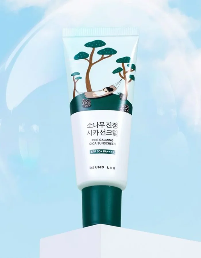 Tube of Round Lab Pine Calming Cica Sunscreen SPF 50 PA against a blue sky background with trees and a hammock illustration.