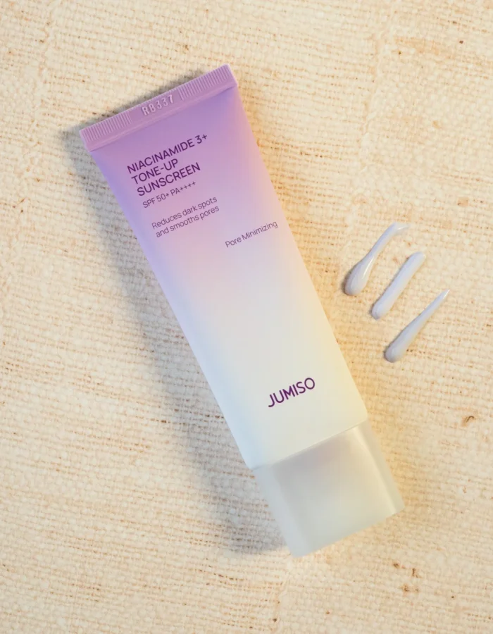 A tube of Niacinamide 3+ Tone-Up Sunscreen SPF 50+ with three small dollops of product beside it on a burlap textured surface.