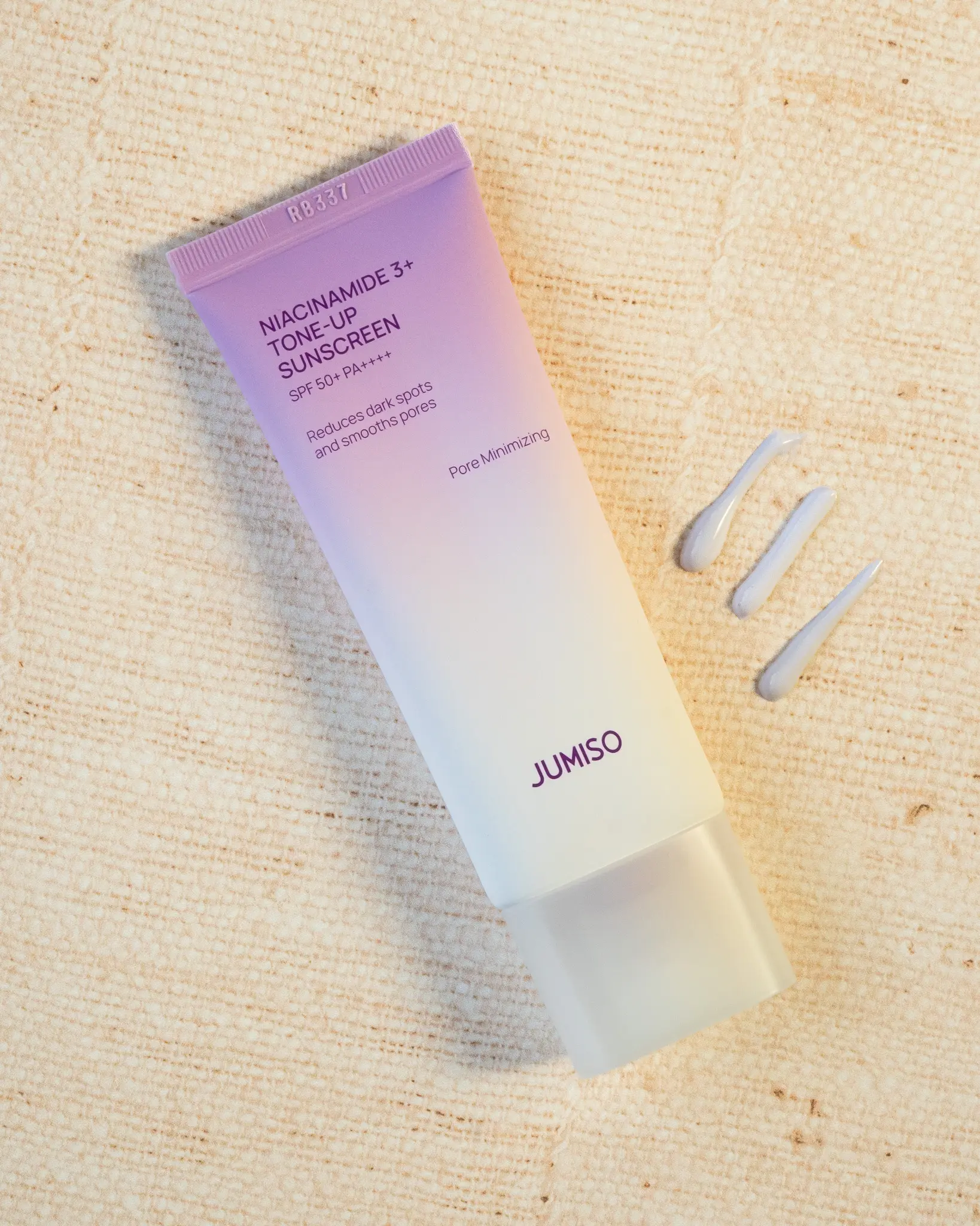 A tube of Niacinamide 3+ Tone-Up Sunscreen SPF 50+ with three small dollops of product beside it on a burlap textured surface.