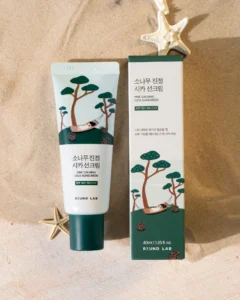 Round Lab Pine Calming Cica sunscreen tube and box on sandy surface, featuring illustrations of trees and a hammock.