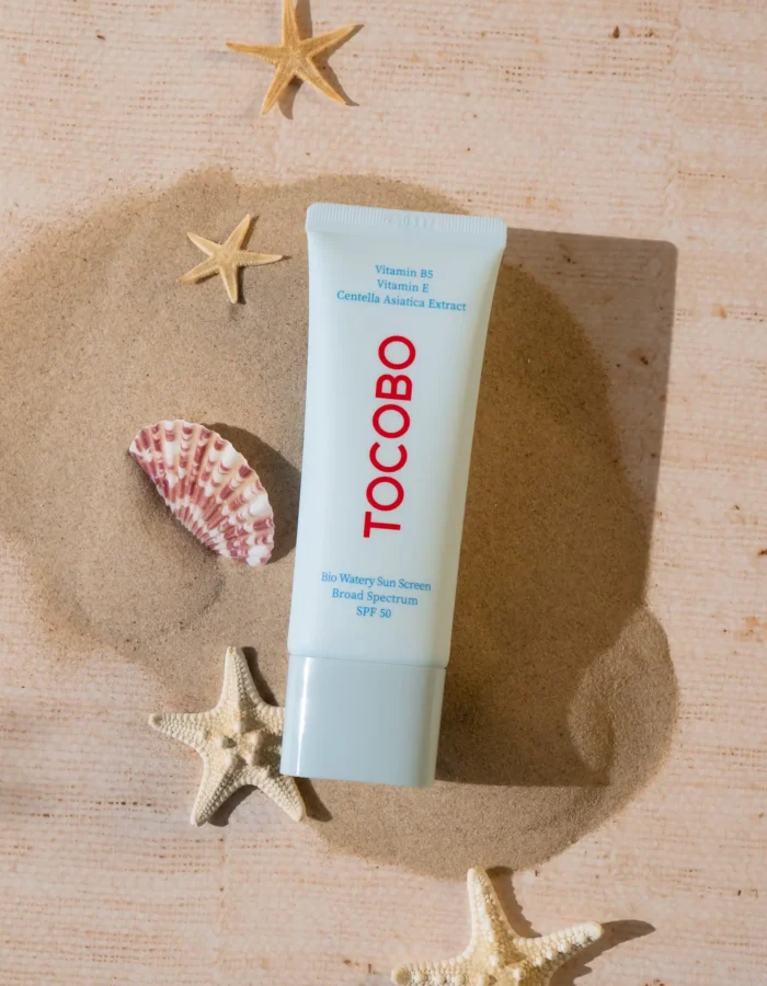 A tube of Tocobo Bio Watery Sun Cream SPF 50 on sandy beach with seashells and starfish in the background.