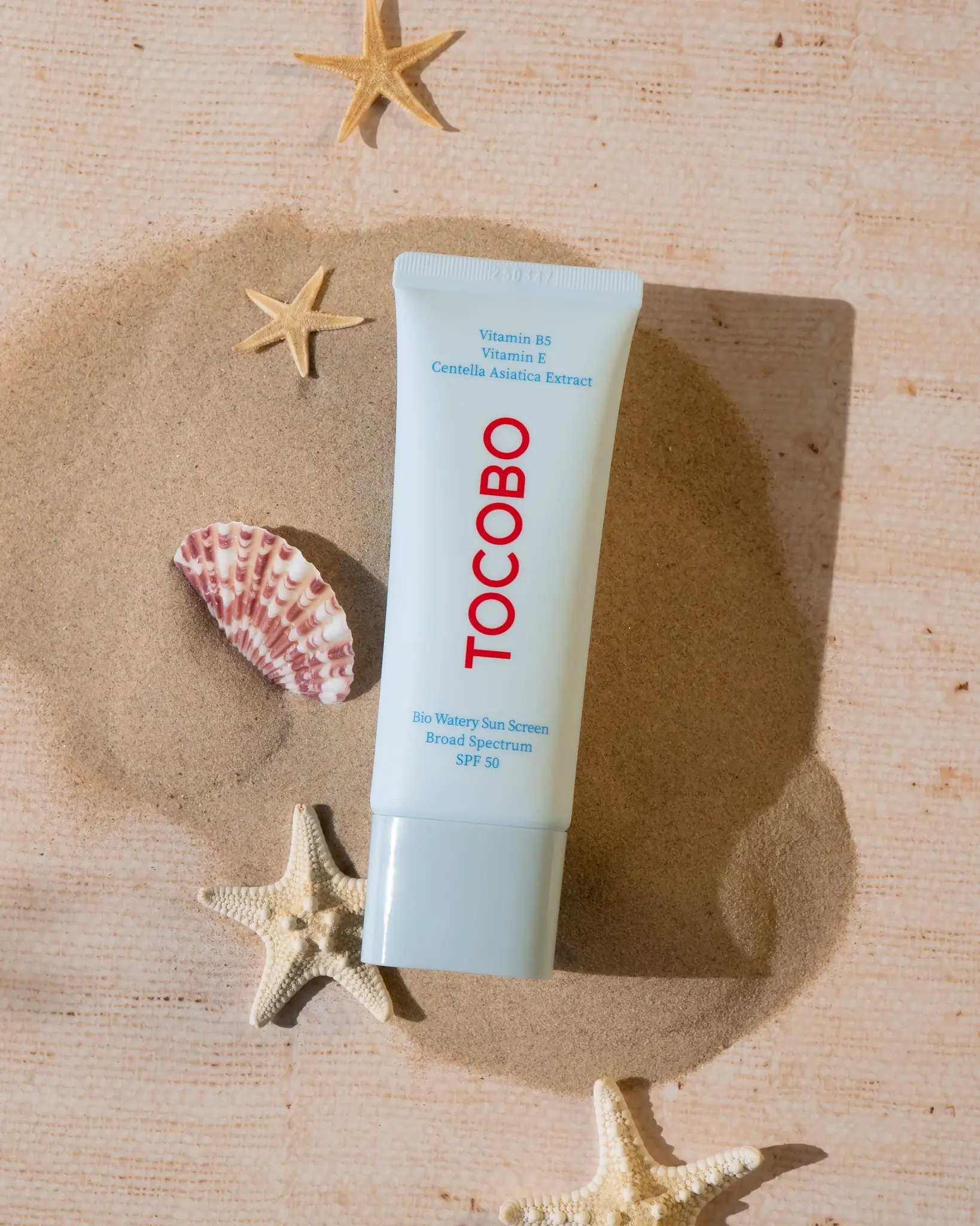 A tube of Tocobo Bio Watery Sun Cream SPF 50 on sandy beach with seashells and starfish in the background.