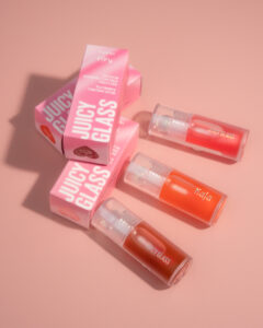 Three Kaja Juicy Glass Lip Oils in clear tubes, with pink packaging, arranged on a soft pink background.