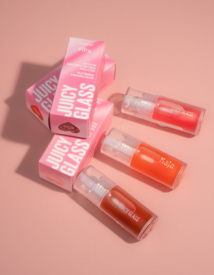 Three Kaja Juicy Glass Lip Oils in clear tubes, with pink packaging, arranged on a soft pink background.