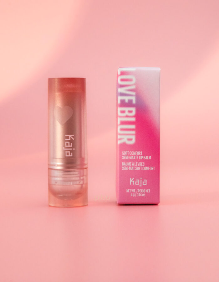 Kaja Love Blur Semi Matte Lip Balm in a clear tube next to its pink packaging on a soft pink background.