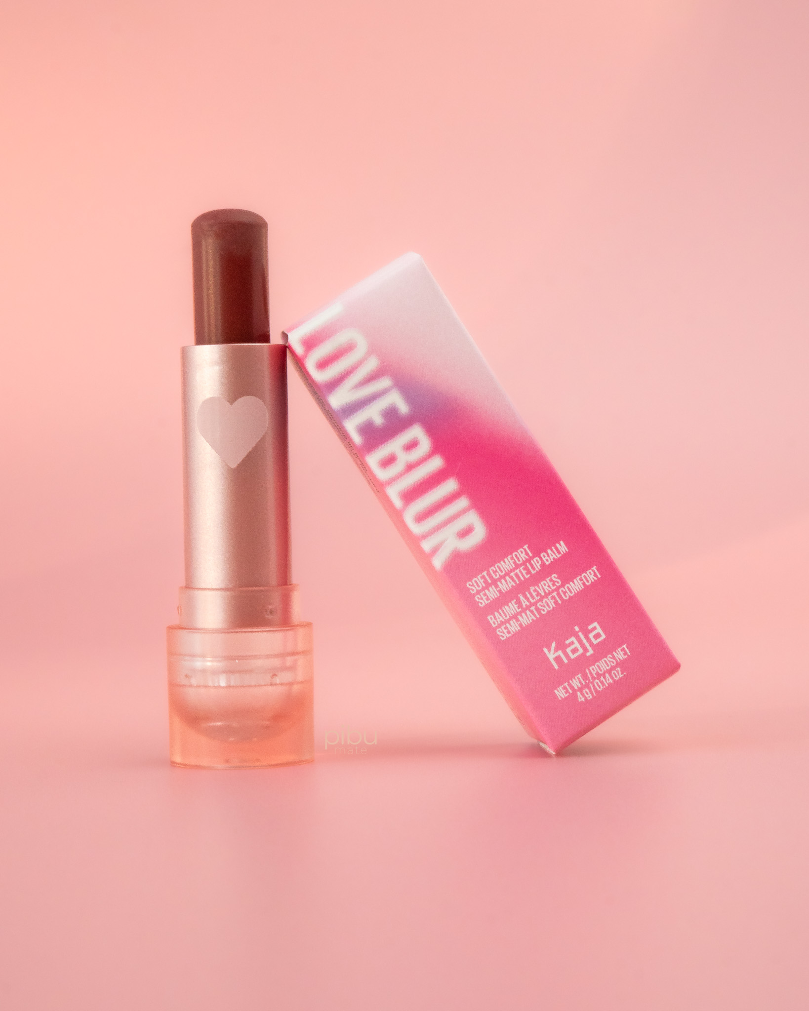A semi-matte lip balm in a tube with a heart design, next to its pink packaging on a soft pink background.