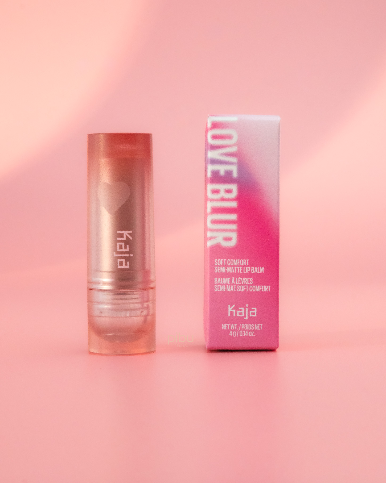 Kaja Love Blur Semi Matte Lip Balm in a clear tube next to its pink packaging on a soft pink background.